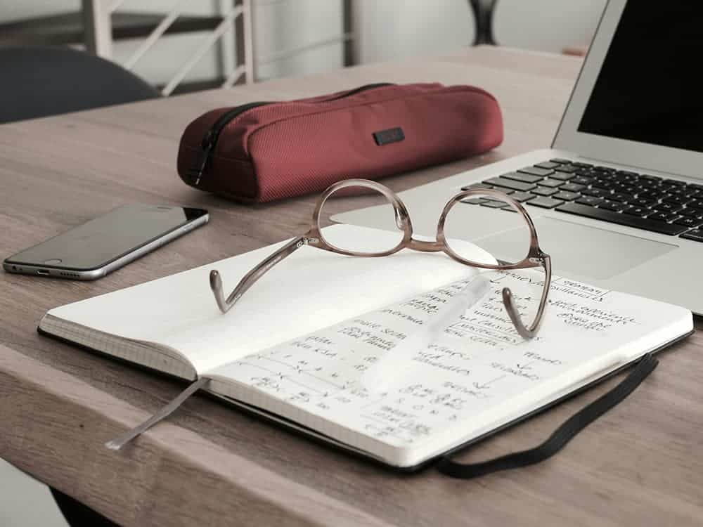 Work desk with open notebook, reading glasses, laptop, and pencil case for productivity.