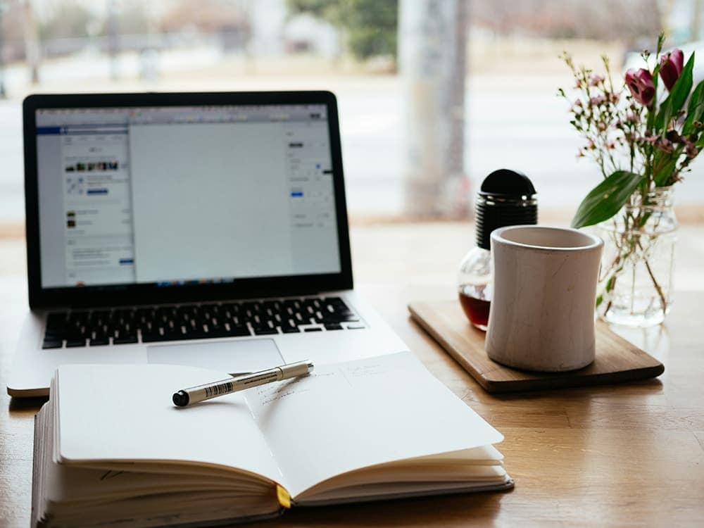 Work desk with open laptop, notebook, pen, and flowers for productivity.